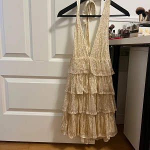 Beige glittrig klänning - Snygg beige klänning med glittriga detaljer och volanger. Den har en djup v-ringning och knytband i nacken. Perfekt för festliga tillfällen.