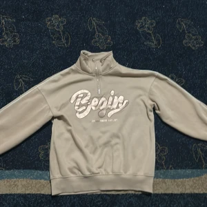 Beige half zip sweatshirt med tryck - Beige sweatshirt med halv dragkedja och hög krage. Framsidan har ett metallic-rosa tryck med texten 'Begin'. Mjuk fleece på insidan och ribbade muddar. Perfekt för chill dagar och snygg till jeans eller leggings.