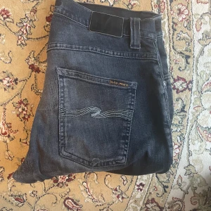 Svarta Nudie Jeans med slitningar - Säljer ett par svarta jeans från Nudie Jeans med snygga slitningar på knäna och klassiska broderade bakfickor. Jeansen har en rak passform och är tillverkade i mjukt bomullstyg. Perfekta för en avslappnad och trendig look.