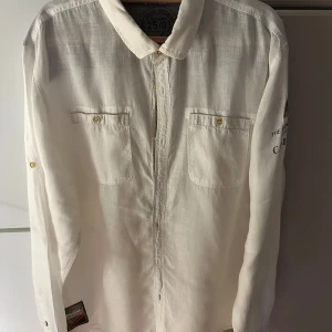 Vit linneskjorta från Camp David - Vit långärmad skjorta från Camp David i linne med broderade detaljer och tryck på ärmen. Skjortan har två bröstfickor med knappar och klassisk krage. Regular fit för en avslappnad men snygg look. Nytt och oanvänd