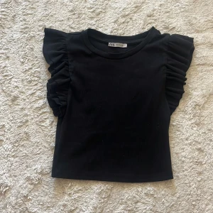 Svart volangtopp från Zara - Supersnygg svart topp från Zara med volangdetaljer på axlarna som ger en cool och trendig look. Toppen är ribbad och har en rund halsringning. Perfekt för dig som vill sticka ut lite extra med dina outfits!