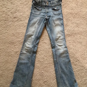 Bootcut jeans från H&M ljusblå - Ljusblå bootcut jeans från H&M med låg midja och justerbar midja. Klassiska fickor fram med lock och knappdetaljer. Tillverkade i bomull, polyester och elastan för en bekväm passform. Perfekta för dig som gillar retrovibbar och snygg siluett.