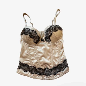 Champagnefärgad satin korsett med spets - Supersnygg korsettopp i champagnefärgad satin med svarta spetsdetaljer över bysten och nedtill. Justerbara axelband och liten rosett.