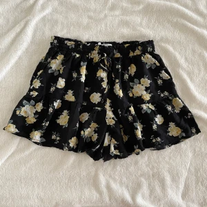 Svarta blommiga shorts från Hollister - Svarta shorts från Hollister med gult och vitt blommönster. De har resår och snörning i midjan för en bekväm passform. Lätta och luftiga, perfekta för varma dagar.