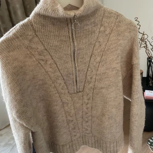 Beige stickad tröja med dragkedja från Bershka - Mysig beige stickad tröja från Bershka med hög krage och halv dragkedja framtill. Tröjan har kabelstickade detaljer längs framsidan och ribbade muddar vid ärmslut och nederkant. Perfekt för sommarkvällar☀️