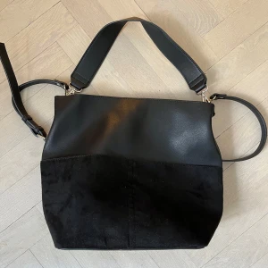 Svart axelväska i skinn och mocka - Säljer en stilren svart axelväska med både slätt skinn och mockadetaljer. Väskan har justerbar axelrem och silverfärgade metalldetaljer. Perfekt för dig som vill ha en enkel men snygg accessoar till vardagslooken.