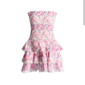 Blommig axelbandslös volangklänning - Supersöt axelbandslös klänning med blommigt mönster i rosa, lila och vitt. Smockad överdel och volanger i kjolen med spetsdetaljer. Perfekt för sommarens alla tillfällen!