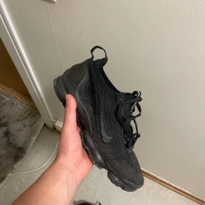 Svarta Nike Air Vapormax sneakers - Säljer ett par helsvarta Nike Air Vapormax sneakers med stickad ovandel och snörning. Skorna har en tydlig Nike-logga på sidan och den klassiska genomskinliga bubbliga sulan. Perfekta för dig som gillar sportig och modern stil.