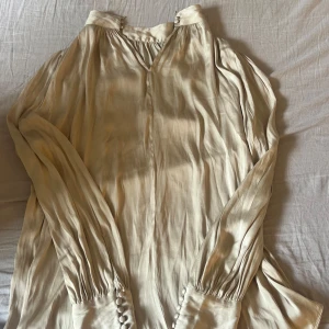 Beige skjorta/blus från H&M - Säljer en beige skjorta/blus i satin från H&M. Blusen har långa ärmar med knappar vid ärmsluten och en lätt v-ringning. Perfekt för dig som vill ha en stilren och tidlös look.