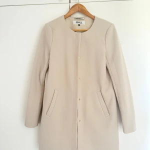 Beige kappa från ONLY - Säljer en stilren beige kappa från ONLY med rak passform och diskreta fickor framtill. Jackan har rund halsringning och stängs med dolda knappar. Perfekt för dig som gillar en clean och klassisk look. Storlek S men passar M också.