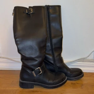 Svarta höga boots med spännen - Säljer ett par svarta höga boots med två silverspännen och dragkedja på sidan. Skorna har en rund tå och platt sula, tillverkade i skinnliknande material. Endast använda fåtal gånger och säljes för att dem blivit för små. Passar storlek 38. 