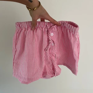 Rosa och vita randiga shorts - Säljer ett par rosa/vita pyjamas shorts med smala ränder. De har elastisk midja och knäppning framtill med knapp och snörning. Perfekta för varma dagar och har en avslappnad passform.