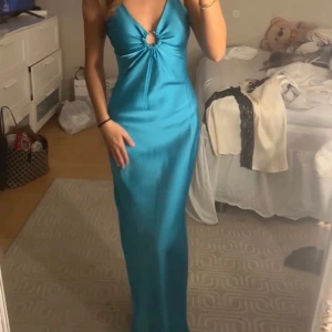 Turkos långklänning med ringdetalj - Säljer en turkos långklänning med djup v-ringning och snygg ringdetalj framtill. Klänningen har smala axelband och är figurnära med ett elegant fall. Perfekt för speciella tillfällen.