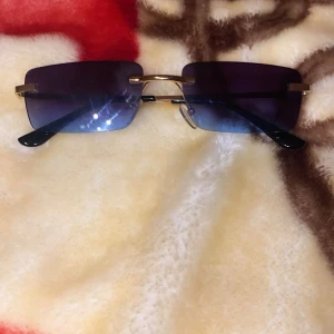 Svarta rektangulära solglasögon med gulddetaljer - Stilrena solglasögon med rektangulära svarta glas och tunna guldfärgade bågar. Glasen är mörka och bågarna är minimalistiska utan ram runt glasen. Perfekta för outfit.