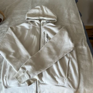 Vit hoodiejacka från Sail Racing - Vit hoodiejacka från Sail Racing med dragkedja framtill och en extra bröstficka med dragkedja. Jackan har huva och broderad logga på bröstet. Mjuk insida och sportig look, perfekt för chill dagar.