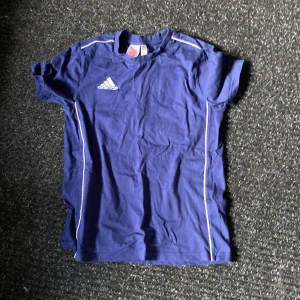 Blå t-shirt från Adidas - Säljer en blå t-shirt från Adidas med vita detaljer och logga på bröstet. T-shirten har korta ärmar och är tillverkad i ett mjukt material, perfekt för träning eller fotboll. Klassisk och sportig stil.