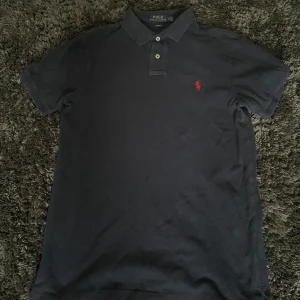 Marinblå pikétröja från Polo Ralph Lauren - Klassisk marinblå pikétröja från Polo Ralph Lauren med röd broderad logga på bröstet. Slim fit-modell med korta ärmar och två knappar vid kragen. Perfekt för en stilren och enkel look.
