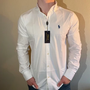 Helt Ny vit skjorta från Polo Ralph Lauren - Klassisk vit skjorta från Polo Ralph Lauren i slim fit-modell. Skjortan har knappar framtill, button down-krage och den ikoniska broderade loggan i marinblått på bröstet. Perfekt för en stilren look.