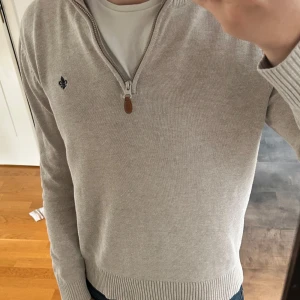 Beige stickad tröja med halv dragkedja från Morris - Säljer en stilren beige stickad tröja från Morris med halv dragkedja och ribbade muddar. Tröjan har en liten broderad logga på bröstet och hög krage. Perfekt för lager-på-lager och passar till jeans eller chinos. Priset kan såklart diskuteras.
