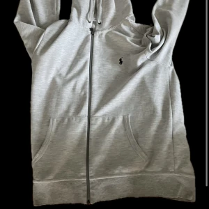 Ljusgrå hoodie från Polo Ralph Lauren - Säljer en stilren ljusgrå hoodie från Polo Ralph Lauren med dragkedja framtill och klassisk logga på bröstet. Tröjan har huva med snörning och två fickor framtill. Perfekt för en avslappnad look. (Den som köper innan 1juli får en pessi max med sig)