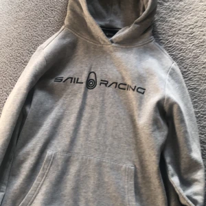 Grå hoodie från Sail Racing - Säljer en stilren grå hoodie från Sail Racing med svart logga på bröstet. Tröjan har huva och en stor magficka framtill. Perfekt för en avslappnad look. Använd bara 2 gånger, den är helt ny kan man säga.