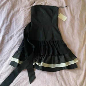 Svart volangklänning med vita detaljer från Zara - Supersöt svart kortklänning från Zara med volanger och vita band längst ner. Axelbandslös med knytband i midjan och kjol med flera lager, bra kvalite. HELT oanvänd och lapp finns kvar. Nypris 60€ (ca 600 kr). Passar perfekt till fest osv💞 Pris går att diskutera vid snabb affär 