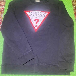 Svart sweatshirt från Guess med logga - Svart sweatshirt från Guess med den klassiska trekantsloggan i rött och vitt framtill. Tröjan har rund halsringning och långa ärmar. Perfekt för en stilren och avslappnad look. Pris kan diskuteras.