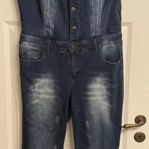 Jeans jumpsuit med knappar - Säljer en blå jeans jumpsuit med knappar framtill och fickor både fram och bak. Snygga sömdetaljer och lätt tvättad look på benen. Perfekt för dig som vill ha en cool och avslappnad stil.