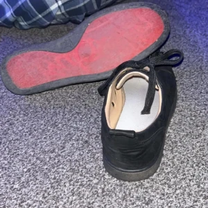Svarta sneakers med nitar och röd sula - Säljer ett par svarta sneakers med svarta nitar på tån och klassisk röd sula. Skorna har snörning och en låg profil, med ovandel i textil och detaljer i syntet. Perfekta för dig som vill sticka ut med en edgy stil.