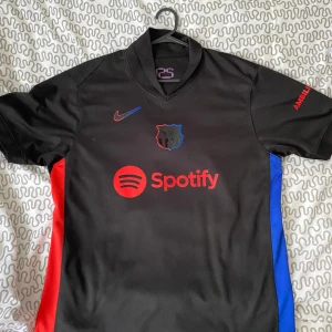 Svart FC Barcelona fotbollströja Nike - Säljer en svart FC Barcelona fotbollströja med röd Nike-logga och Spotify sponsortryck. Tröjan har klubbmärket på bröstet och färgdetaljer i rött och blått på sidorna. Perfekt för dig som älskar Barca!