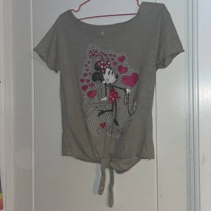 Grå Disney Parks topp med Minnie Mouse-tryck - Grå topp från Disney Parks med stort tryck av Minnie Mouse och rosa hjärtan framtill. Toppen har korta ärmar och knytning nedtill för en trendig look. Perfekt för dig som älskar Disney och vill ha något lekfullt i garderoben.