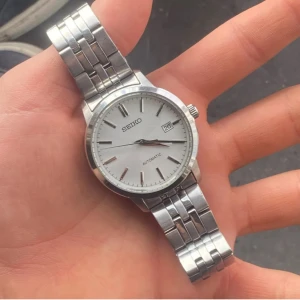Seiko automatisk klocka med datum - Stilren Seiko herrklocka med automatisk urverk och datumvisning. Klockan har en silverfärgad urtavla, markerade index och ett robust armband i rostfritt stål. Baksidan har transparent fönster som visar urverket.