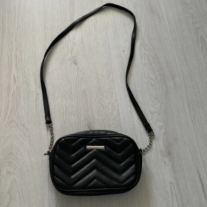 Svart quiltad axelväska med kedjedetaljer - Snygg svart axelväska med quiltat mönster och silverfärgade kedjedetaljer på axelremmen. Väskan har dragkedja upptill och ett innerfack med dragkedja. Perfekt storlek för det viktigaste