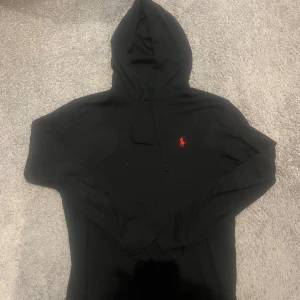 Svart hoodie från Ralph Lauren - Svart hoodie med huva och dragsko från Ralph Lauren. Klassisk design med röd broderad logga på bröstet. Perfekt för en avslappnad och stilren look. Tunn vilket passar sommaren perfekt, pris kan diskuteras vid en snabb affär. 