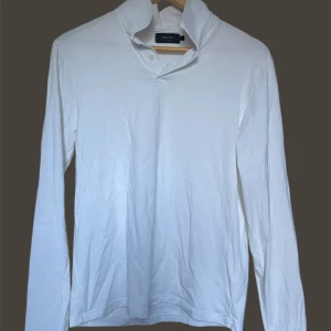 Riley långärmad pikétröja  - Säljer en stilren vit långärmad pikétröja från Riley med klassisk krage och två knappar vid halsen. Perfekt för en clean och enkel look. Passar dig som gillar basic men snygg stil.