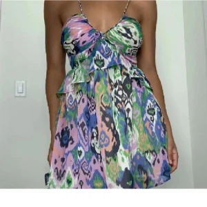 Färgglad kortklänning med mönster - Säljer en färgglad kortklänning med smala axelband och v-ringad byst. Klänningen har ett livfullt mönster i grönt, lila, blått, rosa och vitt samt volangdetalj vid midjan. Perfekt för varma dagar och somriga tillfällen. Skulle säga att den är lite liten I storleken. 