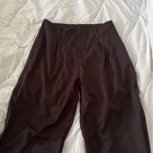 Svart kostymbyxor med pressveck - Säljer ett par svarta kostymbyxor med hög midja och raka ben. Byxorna har klassiska pressveck framtill och stängs med dold dragkedja och hake. Perfekta för en stilren och dressad look.