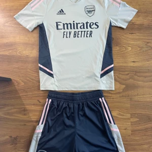 Arsenal träningsset  - Säljer ett Arsenal träningsset från Adidas med kortärmad tröja i ljusgrått och mörkblått samt matchande mörkblå shorts med rosa detaljer. Tröjan har tryckt klubbmärke och sponsorlogga framtill. 