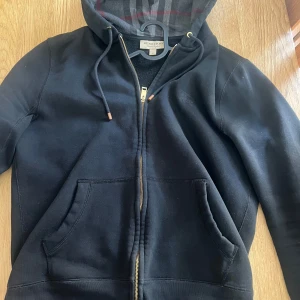 Svart hoodiejacka från Burberry - ÄKTA Svart hoodiejacka från Burberry med dragkedja i guld, klassisk rutigt foder i huvan och två stora fickor framtill. Justerbar huva med snörning och detaljer i metall. Tillverkad i bomull och polyester. Har slutat använda den för den har blivit för liten. Den är i bra kvalite men guld färgen på metall grejer på Sörerna har börjar förvissna. Tröjan är köpt på garmsmarket.