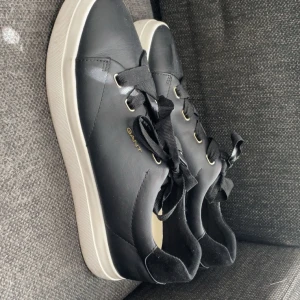 Svarta sneakers från Gant - Snygga svarta sneakers från Gant med vita sulor och svarta snören. Skorna har guldfärgade öljetter och är tillverkade i skinn. Perfekta för en stilren och klassisk look. I modellen ”Avona sneakers i skinn”, nypris 1699kr. Storlek 41. Endast använda 2 gånger!