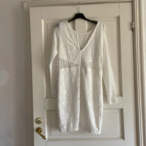 Vit spetsklänning med v-ringning - Säljer en vit kortklänning med lång ärm och djup v-ringning. Klänningen har vackra spetsdetaljer över hela tyget och en figurnära passform. Perfekt för student eller sommarens tillfällen.