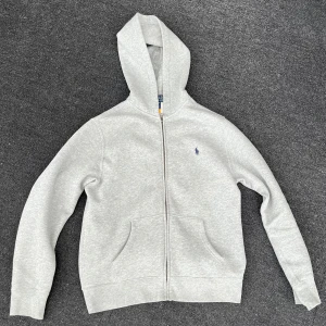 Grå zip hoodie från Polo Ralph Lauren - Säljer en klassisk grå zip hoodie från Polo Ralph Lauren med dragkedja framtill och huva. Tröjan har två fickor och en liten broderad logga på bröstet. Perfekt för en avslappnad stil och enkel att matcha med det mesta. Använd sällan då jag växt ur den fort