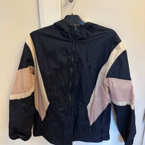 Tommy Hilfiger Sportjacka - Snygg vindjacka med huva från Tommy Hilfiger i svart med beige och ljusrosa detaljer. Jackan har dragkedja framtill och två fickor. Perfekt för blåsiga dagar och har en sportig retrostil.