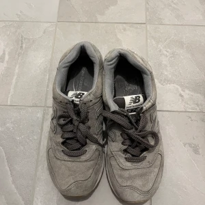 Grå sneakers från New Balance - Säljer ett par klassiska grå sneakers från New Balance med svart logga på plösen. Skorna har snörning och är tillverkade i mocka och syntetmaterial. Perfekta för en avslappnad stil och passar till det mesta.