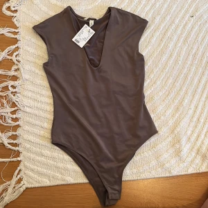 Brun body från hm - Säljer en brun body från H&M i slätt, glansigt material. Den har en stilren design med rund halsringning och en liten slits framtill. Perfekt för dig som gillar minimalistisk stil. Helt ny, aldrig använd💓