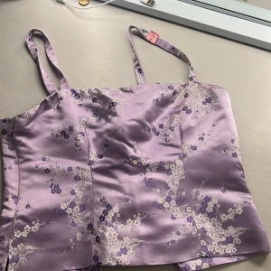 Ljuslila satinlinnetopp från MOJA design - Supersöt ljuslila linnetopp i satin från MOJA design med blommigt mönster i vitt och lila. Toppen har smala axelband och en figurnära passform, perfekt för att lyfta vilken outfit som helst.