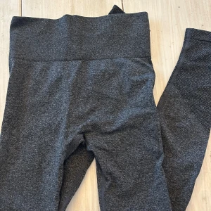 Mörkgrå sömlösa träningstights - Säljer ett par mörkgrå sömlösa tights med hög midja. Tightsen har ribbad linning och diskreta mönsterdetaljer längs sidorna och bak. Perfekta för gymmet eller yoga!