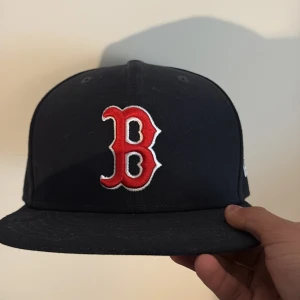 Boston Red Sox keps  - Säljer en svart Boston Red Sox keps från New Era 59FIFTY-kollektionen. Kepsen har en röd och vit broderad B-logga framtill och MLB-logga baktill. Klassisk rak skärm och officiell on-field modell. Färg: Blå och Röd och storlek står på bild 3