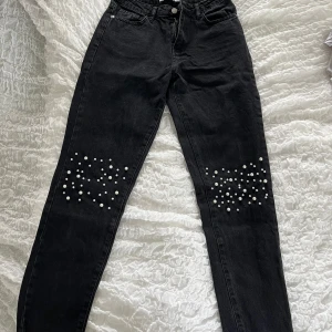Svarta jeans med pärlor från NA-KD - Snygga svarta jeans från NA-KD med raka ben och dekorativa pärlor på båda knäna. Klassisk femficksmodell med dragkedja och knapp framtill. Perfekta för att ge outfiten det lilla extra.