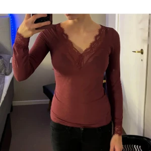 Vinröd långärmad topp med spetsdetaljer - Vinröd långärmad topp med djup v-ringning och spetsdetaljer längs halsringningen. Snygg och feminin passform, perfekt att styla till både jeans och kjol.